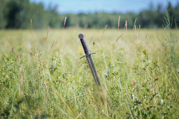 Knight's sword thrust into the ground.  In the field against the background of the forest and the blue sky.  A sunny day of yellow-green steppe. Battlefield. After battle. Crusade, medieval weapons.