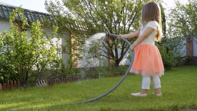 Beautiful little girl in dress water a grass and bushes from a hose