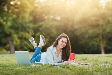 Young girl student  sitting on grass with laptop in front of university and studying homework. She...
