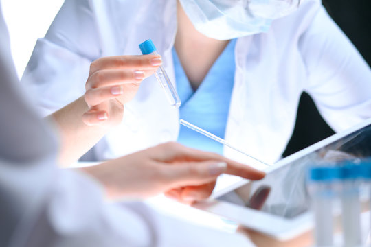 Closeup of scientific research team with clear solution in laboratory. Blonde female chemist holds test tube of glass while her colleague checks results with tablet pc. Blood test, medicine or