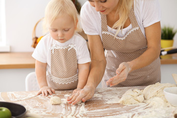 Little girl and her blonde mom in beige aprons  playing and laughing while kneading the dough in the kitchen. Homemade pastry for bread, pizza or bake cookies