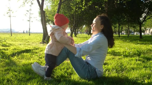 Mother and baby are playing in the parken sunset. Mum lies on the grass and picks up baby feet above her head