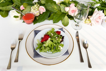 Place setting including a light salad with strawberries on white plates with a table floral arrangement in the background.