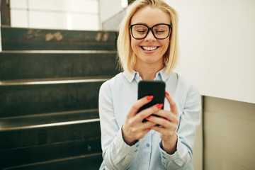 Smiling young businesswoman sitting on office stairs sending tex