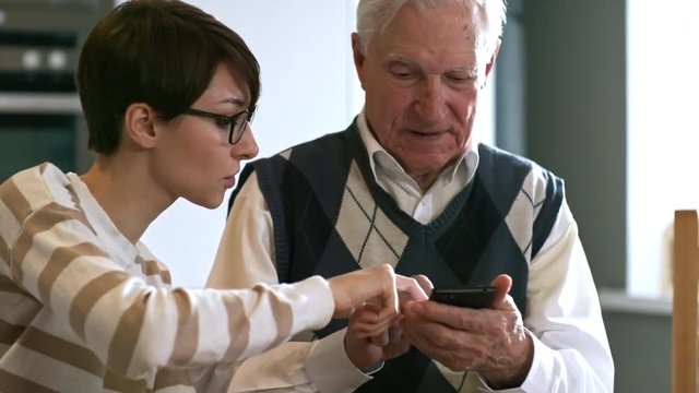 Tilt down of young woman in glasses teaching senior man how to use smartphone