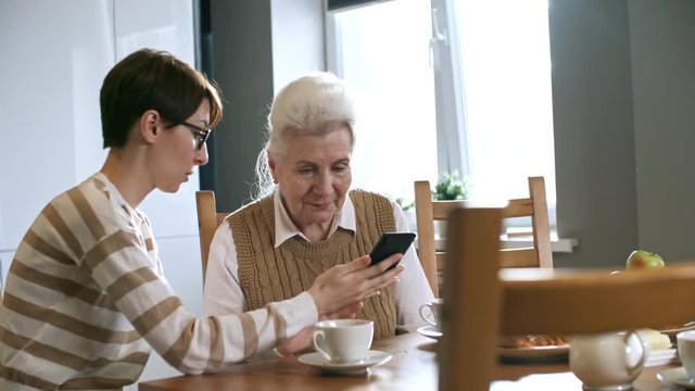 Zoom out shot of young woman in glasses showing smartphone to senior lady sitting at kitchen table, then explaining her how to use it