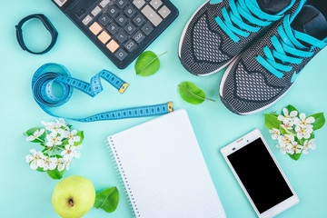 Fitness, healthy and active lifestyles concept. Sport shoes, smartphone, tape, calculator, fitness bracelet and green apple on green or blue background with copy space. Top view