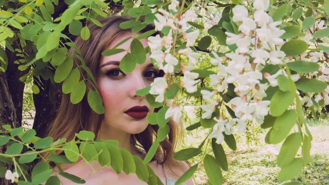 Beautiful young woman surrounded by flowers spring blossom. Girl with a beautiful face and a gentle make-up near the blossoming acacia tree. Girl's look through a blossoming tree.
