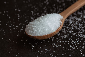 Natural purified sugar or bleach sugar on wood spoon on black granite table in close up view macro concept with copy space. Ingredient prepared for cooking or bakery. Unhealthy seasonings for diet.