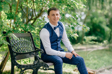 Obraz premium Portrait of business man in suit sitting in park on bench and smiling. The guy is getting ready for the wedding day, waiting for his bride.