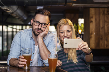 Young couple taking selfie by mobile phone on local pub and she smile while he is boring
