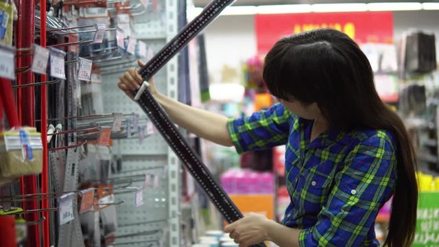 A young woman in a blue checkered shirt chooses and buys a large iron ruler with an angle in the supermarket in the building materials department.