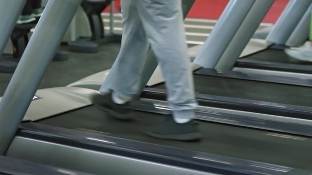 Low-section shot of unrecognizable man in sweatpants and sneakers walking on treadmill in gym