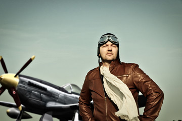 Portrait of a vintage pilot with leather cap, scarf and aviator glasses in front of a historic airplane - Portrait of a man in historical pilot clothing - obrazy, fototapety, plakaty