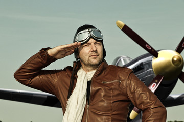 Portrait of a vintage pilot with leather cap, scarf and aviator glasses salutes in front of a...