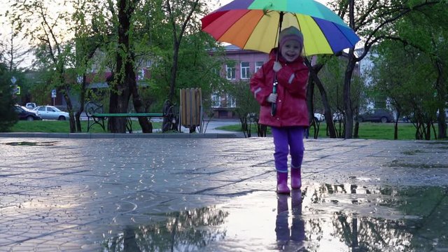 A nice little girl with a colorful umbrella running through the puddles in the park at sunset. The child smiles and enjoys the fun, slow motion.