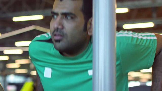 Tilt up of focused Arab man in shirt doing triceps dip exercise in gym while doing workout