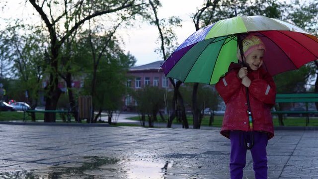 A nice little girl in a red raincoat is standing by a puddle in the park at sunset. Drops of rain flow down a multi-colored umbrella. The child smiles and enjoys the fun.