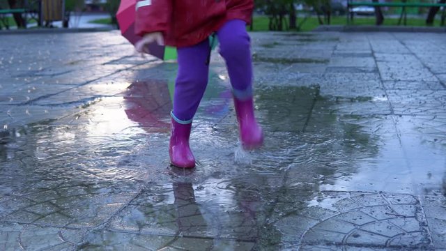 A cute little girl in a red raincoat jumping over a puddle in the park at sunset. The child smiles and enjoys the fun.