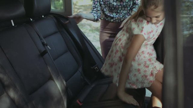 Beautiful caring mother fastening cute elementary age girl with safety seat belt in the back seat of the car while family going on summer vacation road trip. Child buckles up in auto during journey.