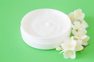 Fototapeta na wymiar Opened jar of natural herbal cream for women on green background. Care about clean and soft face, hands, legs and body skin. Beautiful jasmine blossoms. Fresh, white flowers. Close up.