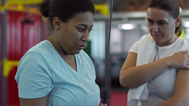 Tilt up of middle-aged woman with towel giving advice to black female friend doing triceps pushdown exercise in gym