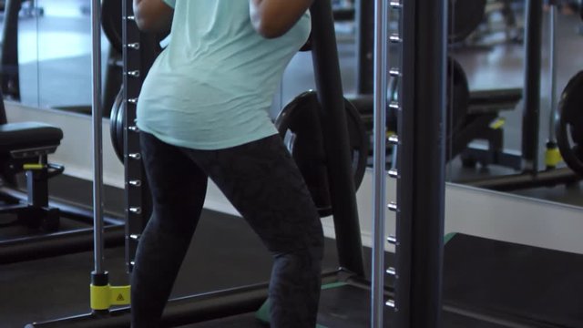 Tilt up of unrecognizable black woman in sportswear doing squats with empty barbell in smith machine