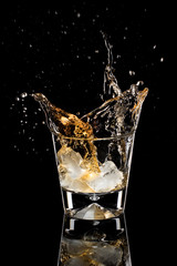 Pieces of ice fall into a glass with whiskey. A photo on a black background.