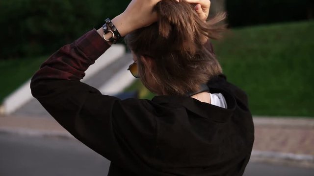 Traking footage of a young girl walking outdoors on the street or park. Cheerful and happy. Making a ponytail on her short fair hair, wearing sunglasses and put on the headphones