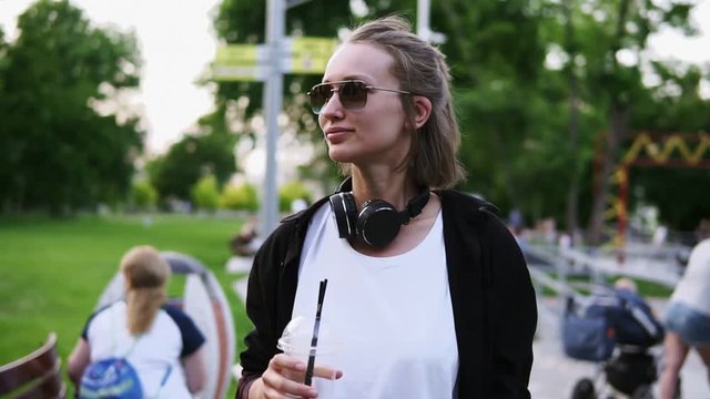 Front traking footage of a young, pretty girl walking through the park. With headphones on her neck and stylish sunglasses. Leisure time