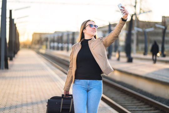 Woman taking selfie with mobile phone in train platform at station. Smiling and happy lady taking photo of herself with smartphone. Traveler and tourist arrived to destination.