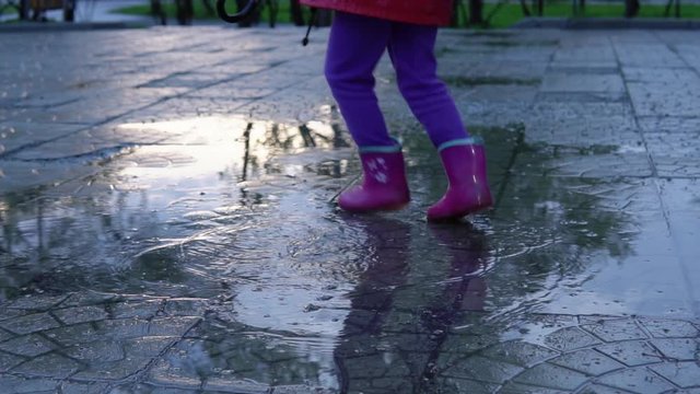 A cute little girl in a red raincoat jumping over a puddle in the park at sunset. The child smiles and enjoys the fun, slow motion.