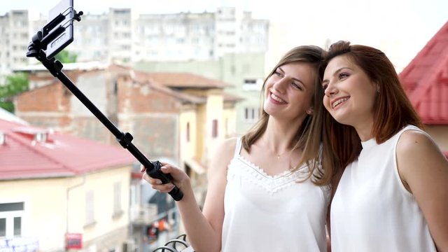 Two best friends smiling and laughing while taking a selfie photo with a selfie stick on the balcony. Slow motion