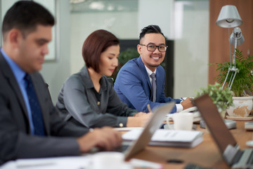 Working meeting at spacious boardroom: multi-ethnic group of managers in formalwear sitting at wooden table and brainstorming on ambitious project, handsome Asian man posing for photography