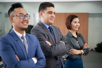 Multi-ethnic team of smiling managers looking away while standing in raw at spacious open plan office with arms crossed, group portrait shot