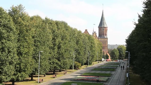 Cathedral of Kant in Kaliningrad, the alley with unrecognizable pedestrians. Old medieval castle at summer windy day. An ancient сatholic gothic tower with a clock.