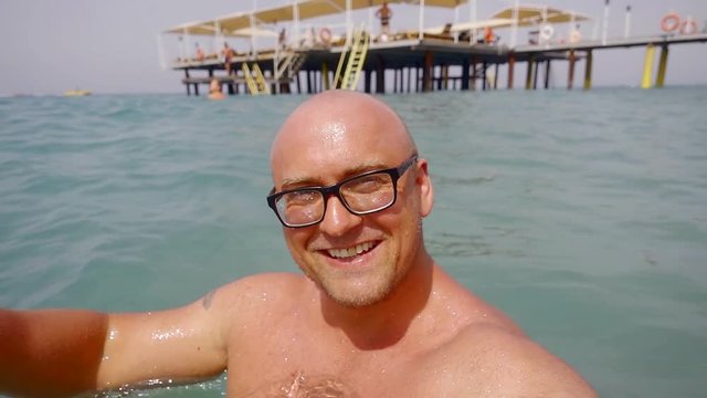 cheerful muscular bald man at the resort is swimming in the sea next to the pier with glasses and a white straw hat