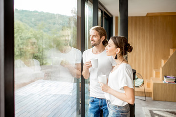 Young and happy couple dressed in white shirts standing together with cups near the window at home - Powered by Adobe