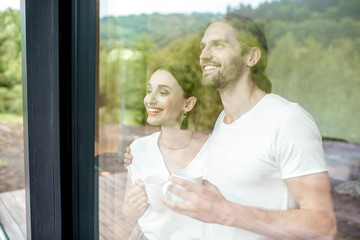 Young and lovely couple standing together looking outside the window at home, view through the...