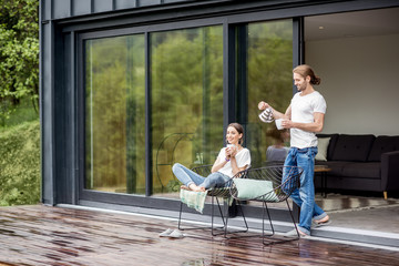 Young couple sitting with cups on the terrace of the modern house enjoying beautiful view outdoors - obrazy, fototapety, plakaty