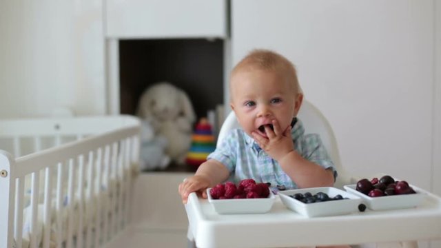 Adorable little baby boy, eating fresh fruits at home, sitting in baby chair in kids room