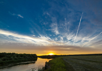 The sun behind the clouds at sunset over the river and meadow.