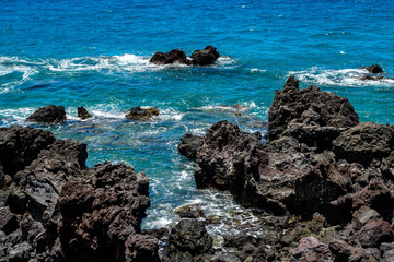 Fototapeta na wymiar Bright blue Pacific Ocean with a rough black lava rock shoreline and crashing waves, Hawaii 