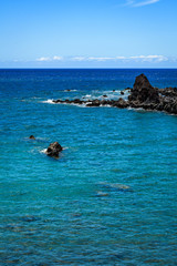 Obraz premium Bright blue Pacific Ocean with a rough black lava rock shoreline and crashing waves, and blue a sky with white clouds in the background, Hawaii 