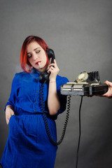 A girl with red hair in a blue dress with a cigarette rings an old phone