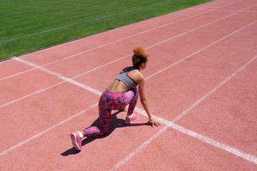 A tanned woman or African American girl prepares to run on the white starting line of the pink treadmill next to the green grass on the football field