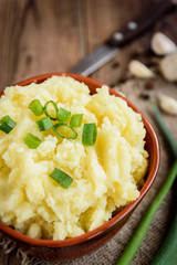 Mashed potatoes in bowl on wooden table with spring green onion and garlic.