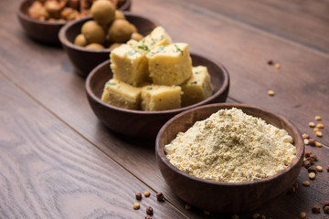 Chick pea flour or Besan powder in a ceramic or wooden bowl along with Gujrati Dhokla snack, onion bajji or bhaji and sweet laddu or laddoo