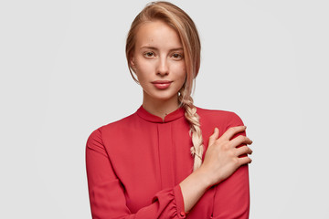 Photo of serious delighted female with long plait, keeps hand on shoulder, wears red elegant...
