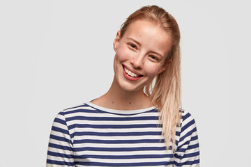 Happy female student with broad smile, has light hair combed in pony tail, recieves positive news, dressed in striped casual jacket, glad to meet with best friend, isolated on white background